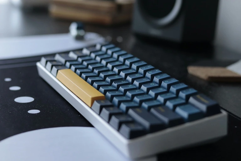 Compact mechanical keyboard with a yellow spacebar beside a desktop speaker